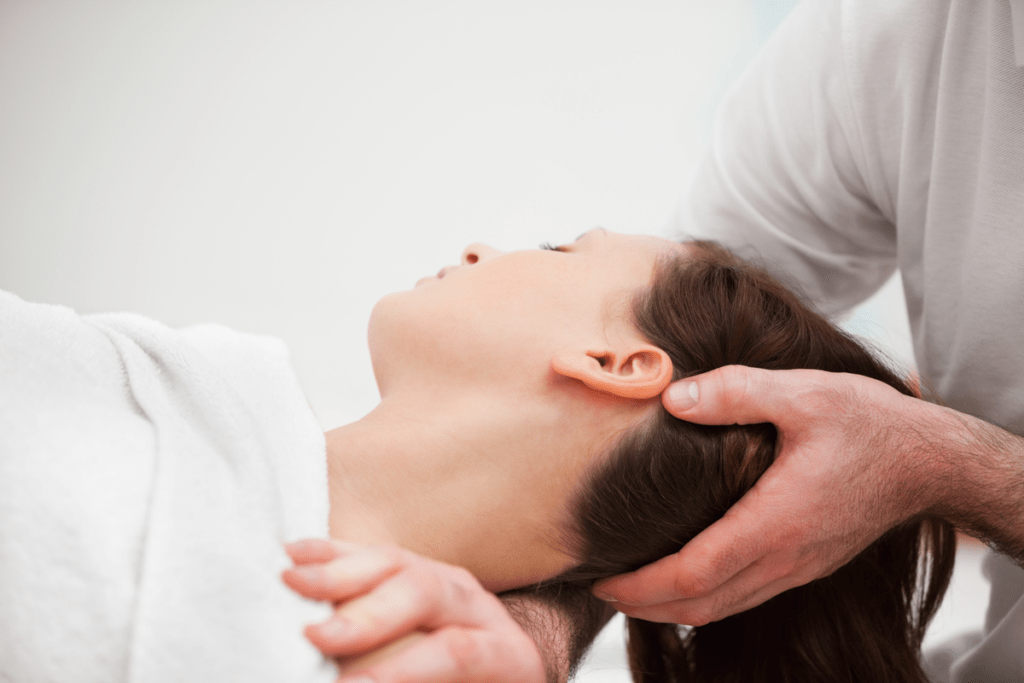 En person som får en nackmassage medan hon ligger ner med en vit handduk draperad över sina axlar. En annan persons händer, som antas vara en massageterapeuts, stödjer försiktigt deras huvud och nacke, vilket illustrerar en terapeutisk eller avslappningssession.